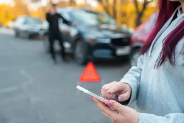 A female with pink nails uses a smartphone near a road with a warning triangle, while a person stands by a car met with an accident