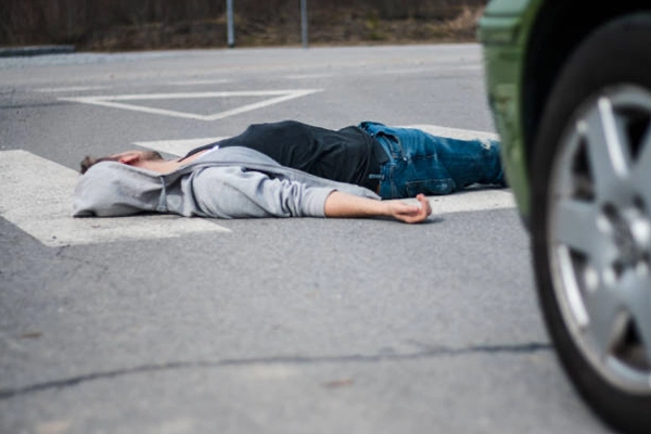 A person wearing a hoodie lies unconscious on a crosswalk, with a car approaching nearby, suggesting a serious accident.
