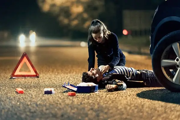 A woman is providing assistance to an injured man lying on a dark road, with a warning triangle and emergency supplies nearby.