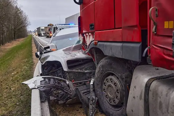 A damaged car crushed against a guardrail by a large red truck on a roadside, with emergency lights flashing in the background.